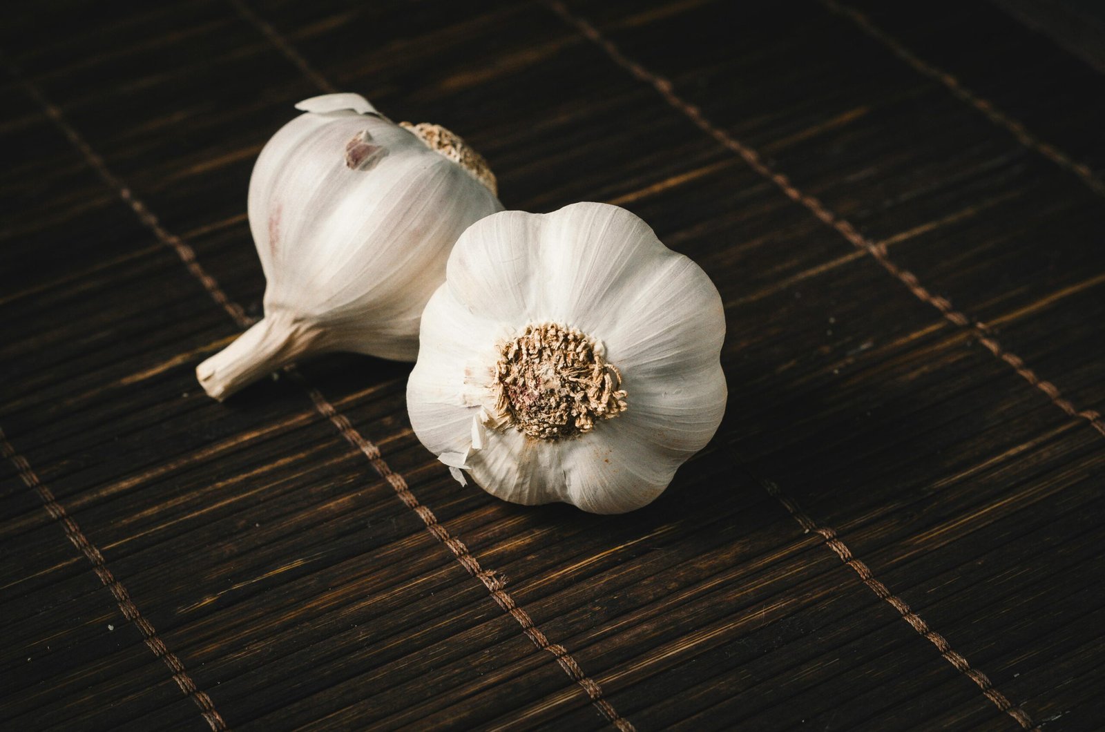 Two fresh garlic bulbs on a dark wooden mat, highlighting aromatic culinary ingredients.